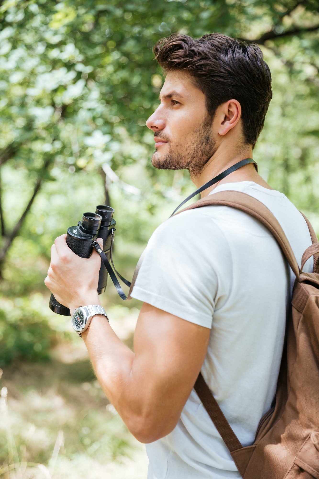 Pensive young man with backpack holding binoculars in forest
