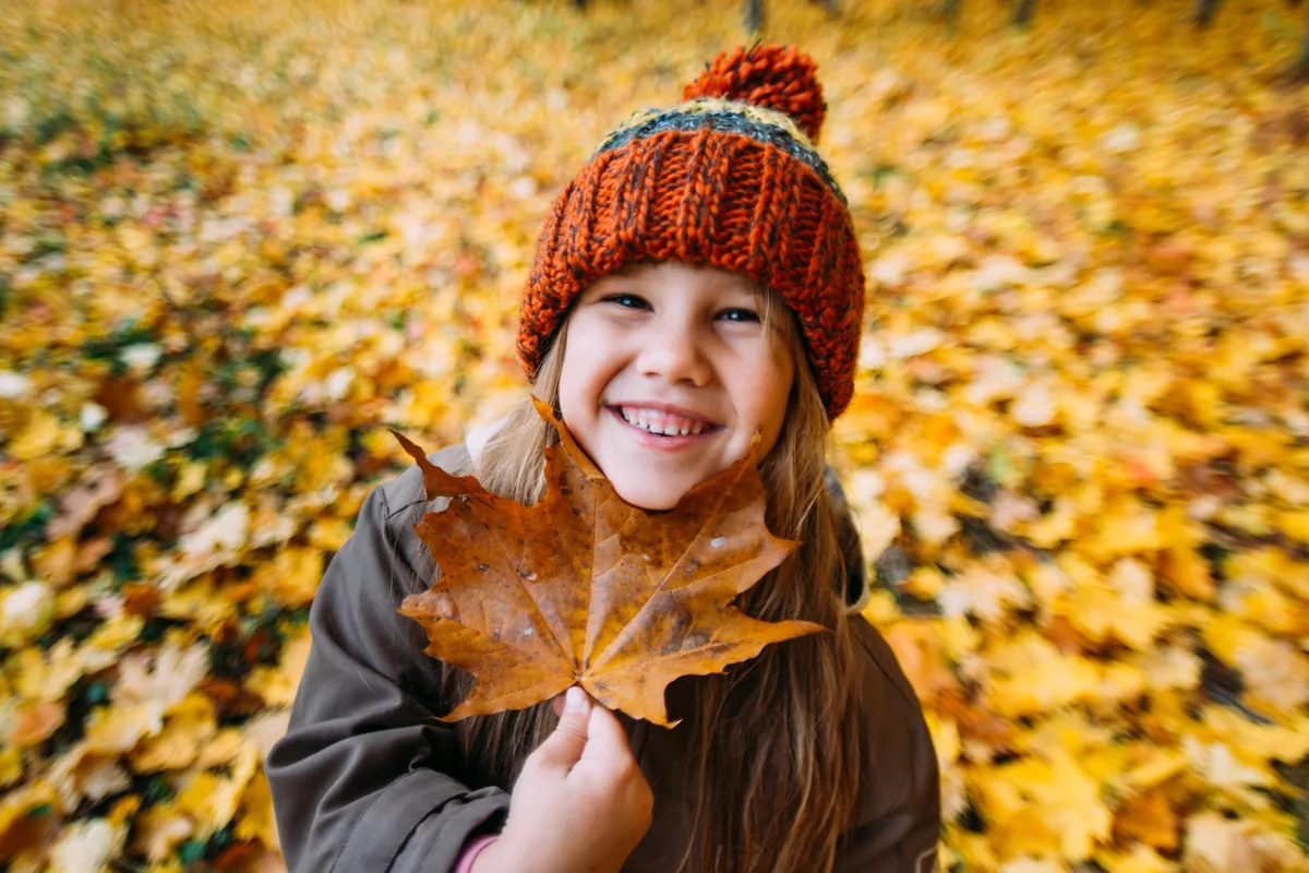 Smiling girl in a knitted hat holding a large maple leaf in front of autumn foliage.