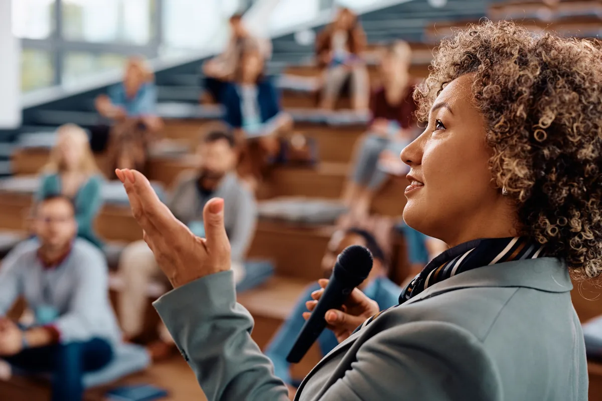 Presenter gesturing with one hand while speaking into a microphone to a seated audience.