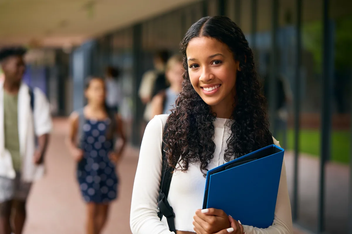 Smiling student with a backpack holding a blue binder on a campus walkway.