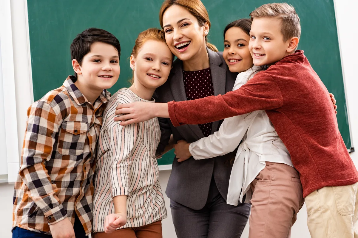Teacher smiling as pupils gather around her for a group hug in front of a chalkboard.