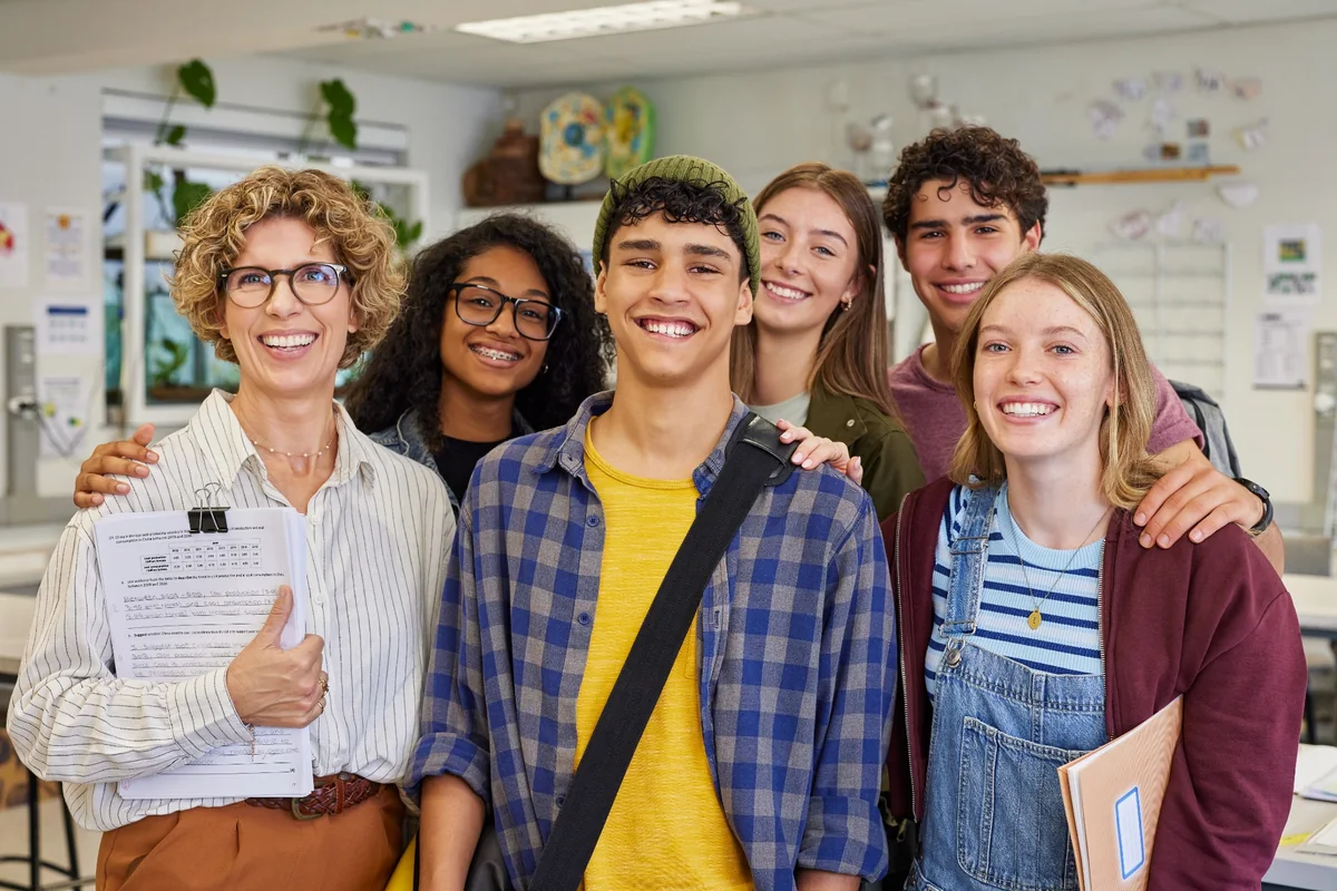 Teacher and smiling teenage students standing together in a classroom with notebooks and papers.