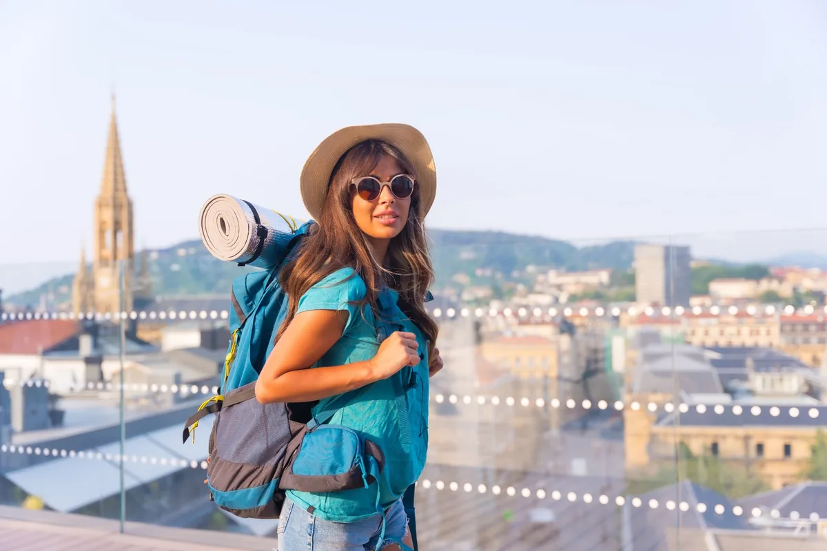 Woman wearing a hat and sunglasses with a backpack overlooking a riverside city.