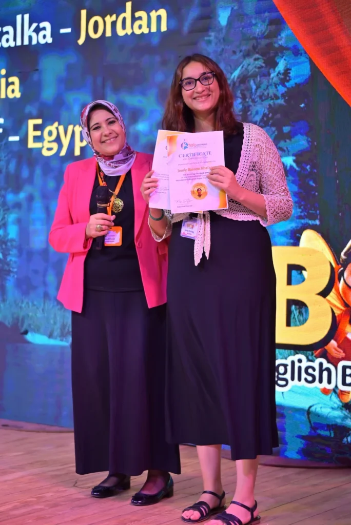 Woman wearing glasses holding a certificate beside a presenter on the WEB Olympiad stage.