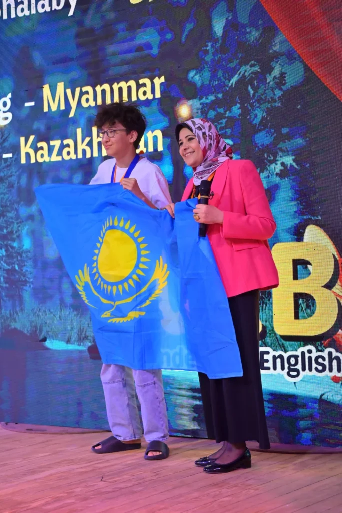 Participant holding the Kazakhstan flag beside a presenter on the WEB Olympiad stage.