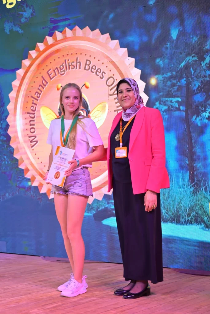 Student wearing a medal and holding a certificate beside a presenter on the WEB Olympiad stage.