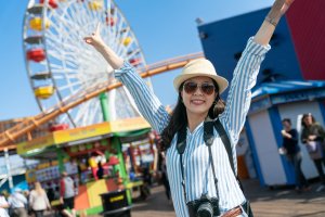 happy stylish asian Japanese female visitor on holiday at amusement park looking at camera with raised arms and victory hand signs on background of a Ferris Wheel